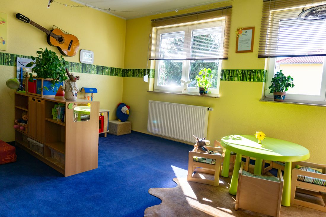 Sonniges Spielzimmer mit grünem Kindertisch, kleinen Holzstühlen, Spielregal und Gitarre an der Wand