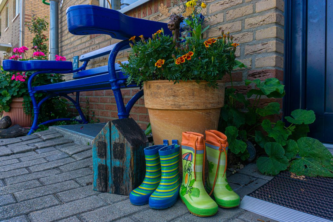 Bunte Kindergummistiefel vor der Haustür neben Blumentopf und blauer Bank